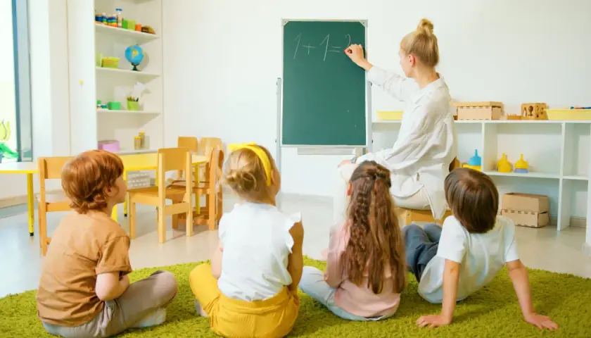 Nauczyciel wychowania przedszkolnego pisze na tablicy "1+1=2". Dzieci siedzą na dywanie i uważnie słuchają.