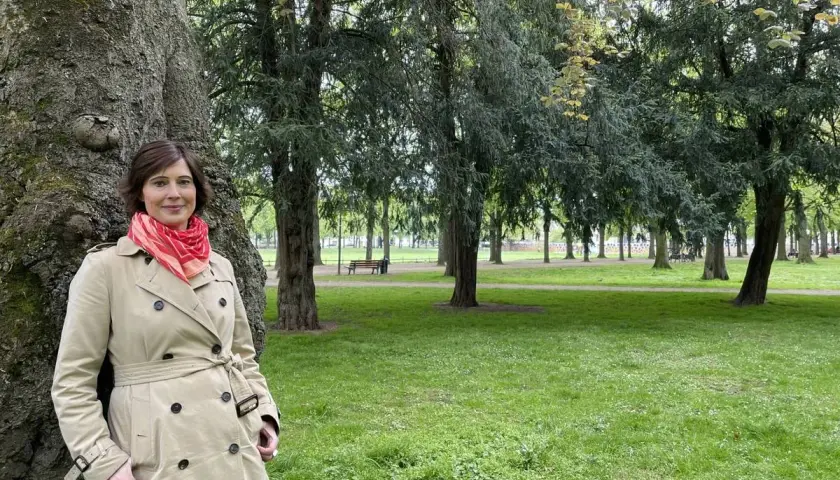 Kristina Hartenbach lehnt an einem Baum im Park. Sie trägt einen beigen Trenchcoat und einen roten Schal.