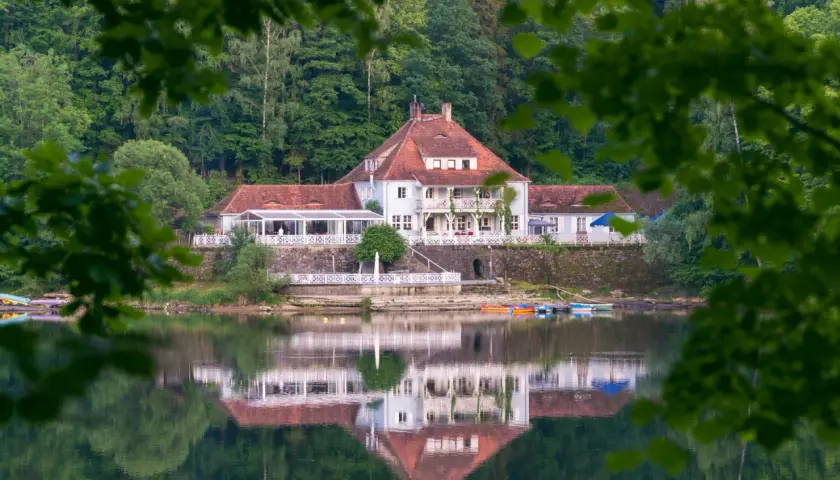 Biały hotel z czerwonym dachem nad spokojnym jeziorem, odbijający się w wodzie.