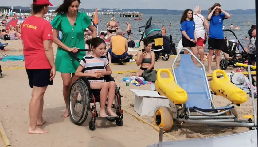 Na plaży dla niepełnosprawnych ratowniczka pomaga dziewczynce na wózku inwalidzkim. W tle widać innych plażowiczów i morze.