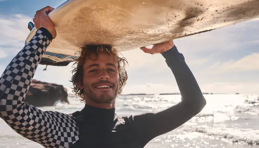 Lächelnder Surfer mit Wellen im Haar trägt sein Brett über dem Kopf. Sein Neoprenanzug hat ein cooles Schachbrettmuster.