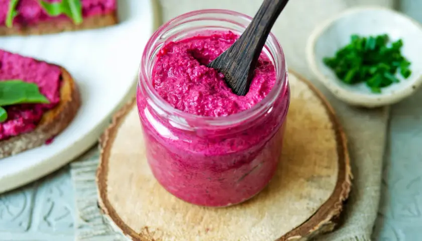 Glas mit cremigem, veganem Rote Bete Aufstrich, daneben Brot mit Aufstrich und Schnittlauch.