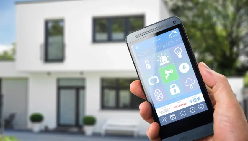 Dłoń trzyma smartfon z aplikacją do **instalacji inteligentnego domu**. Na ekranie widoczne ikony sterowania oświetleniem, ogrzewaniem, kamerami i innymi funkcjami.