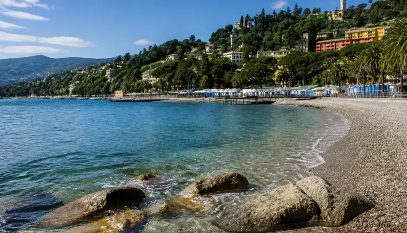 Plage de galets à Rapallo, avec des cabines bleues et blanches alignées, des maisons colorées sur la colline verdoyante et une mer bleue étincelante.