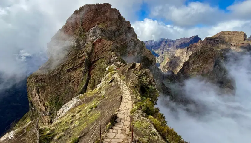 A stone path winds along a narrow mountain ridge, offering what is likely the best hiking in Portugal. Clouds drift through the dramatic, rugged peaks.