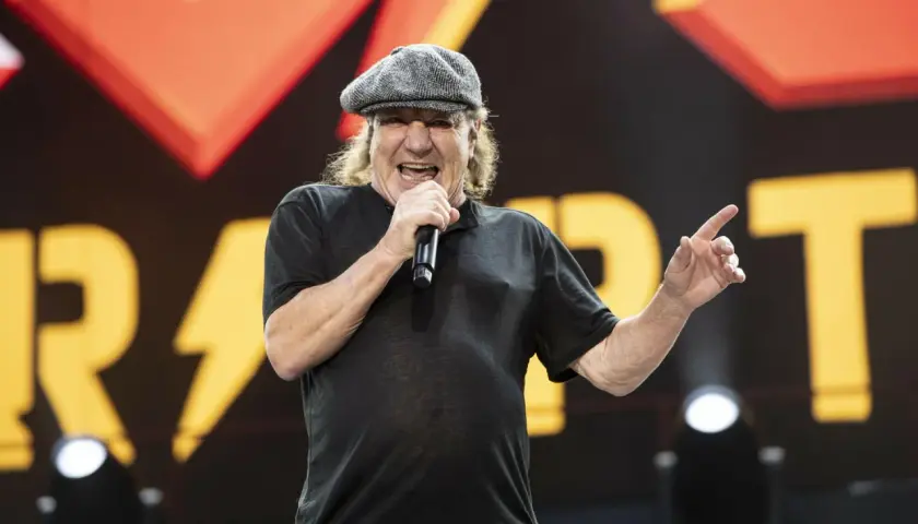 Brian Johnson young, w czapce i czarnej koszulce, śpiewa do mikrofonu na tle sceny z logo zespołu.
