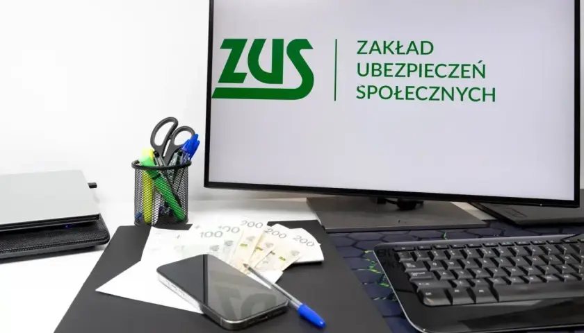 Na biurku leżą pieniądze, telefon i długopis. Na monitorze widnieje logo ZUS, symbolizujące składkę ZUS.