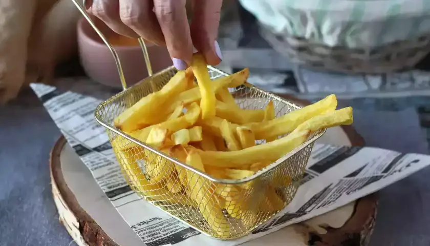 Chrupiące złote frytki w metalowym koszyku, podane na gazecie, ręka sięga po jedną.