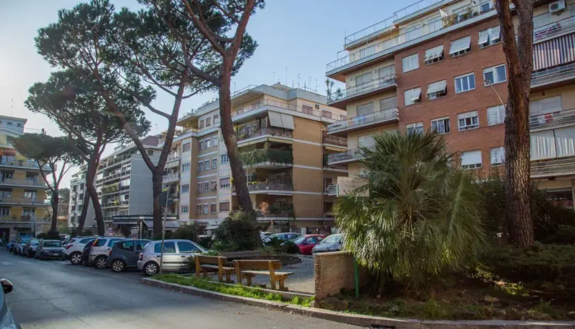 Edifici residenziali e pini marittimi nel quartiere Balduina a Roma. Auto parcheggiate lungo la strada, panchine e vegetazione rigogliosa.