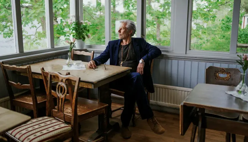 Carl-Johan Vallgren, med sin fru vid sin sida, sitter vid ett bord med utsikt över en grönskande trädgård.