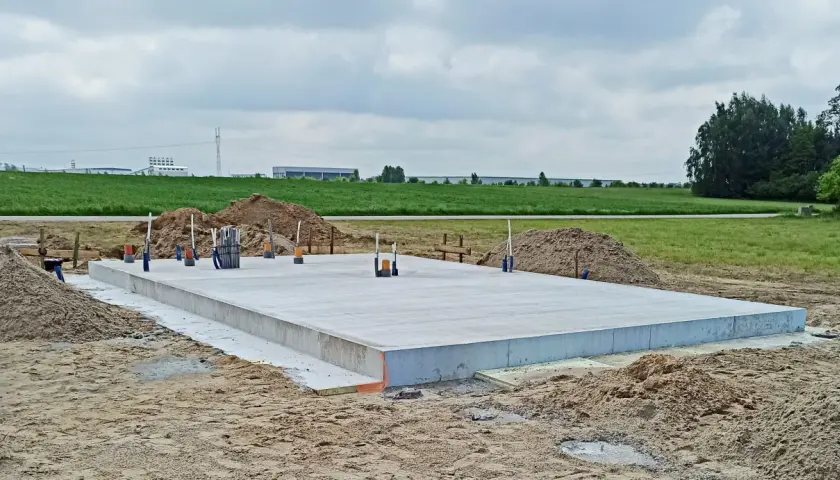 Gotowa płyta fundamentowa na budowie, otoczona piaskiem i zielenią. W tle widać budynki przemysłowe.