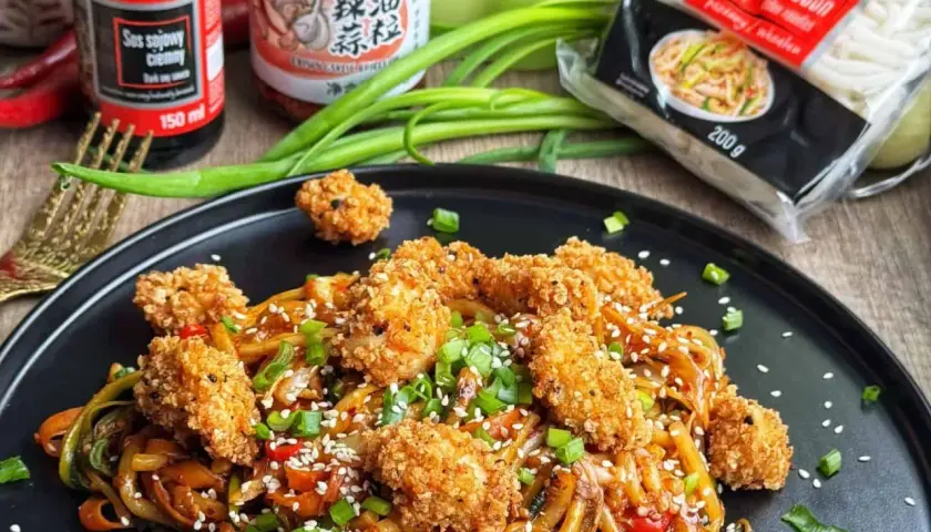 Pyszny makaron udon z kurczakiem w panierce, posypany sezamem i szczypiorkiem. Obok sos sojowy i opakowanie makaronu.