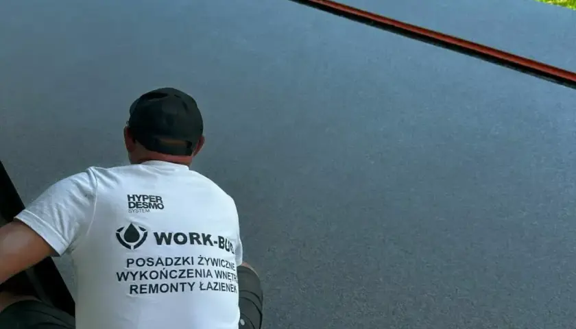 Pracownik w czapce i koszulce z logo firmy wykonuje posadzki żywiczne w garażu.