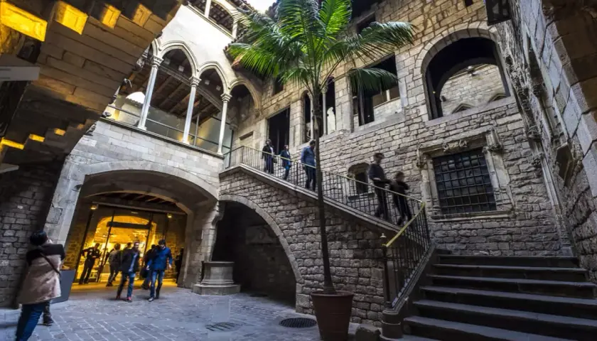 Un patio interior de piedra en el Born, con una palmera y escaleras que suben. Gente pasea por el lugar.