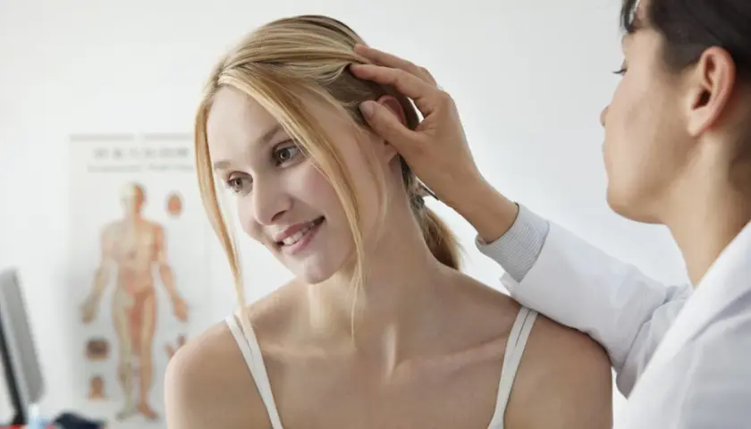 Dermatolog bada skórę głowy młodej kobiety. W tle wisi plakat anatomiczny.