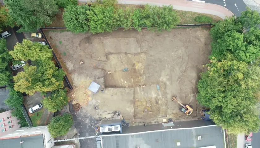 Wykop archeologiczny na placu budowy, gdzie prowadzone są badania przed pozwoleniem na budowę. Widoczny koparka i ślady dawnej zabudowy.