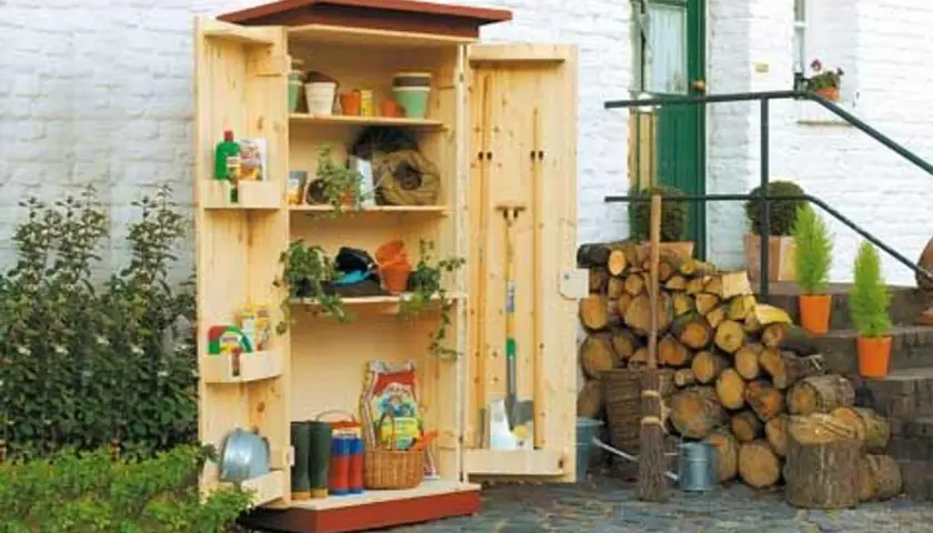 Ein selbstgebauter Gartenschrank aus Holz, gefüllt mit Gartenutensilien wie Töpfen, Werkzeugen und Gummistiefeln. Daneben liegt aufgeschichtetes Brennholz.