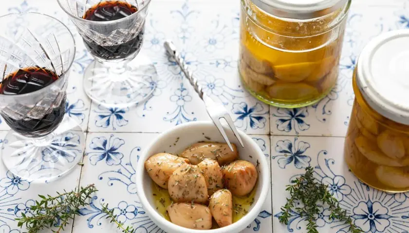 Ail confit au four, tendre et parfumé, servi dans un bol avec des herbes. Deux verres de vin rouge et des bocaux d'ail confit complètent la scène.