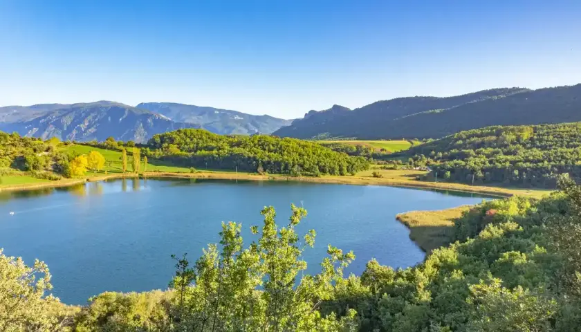 Paisaje sereno en la conca de dalt: un lago azul rodeado de colinas verdes y montañas bajo un cielo despejado.