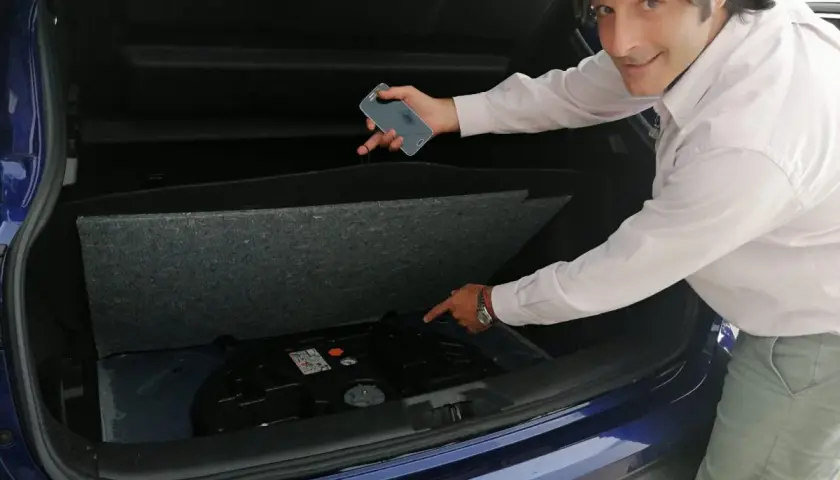 Hombre señala el espacio vacío en el maletero de un coche azul, donde debería estar la rueda de repuesto. "He pinchado y no tengo rueda de repuesto", parece decir.