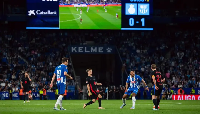 Piłkarze na boisku, jeden z nich w czarnej koszulce z numerem 19, próbuje odebrać piłkę. To przykład taktyki "co to pressing" w akcji. Wynik 0-1 na tablicy.
