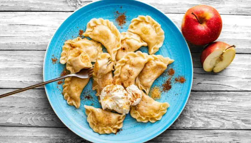 Pyszne pierogi z jabłkami, posypane cynamonem, na niebieskim talerzu, obok jabłka.