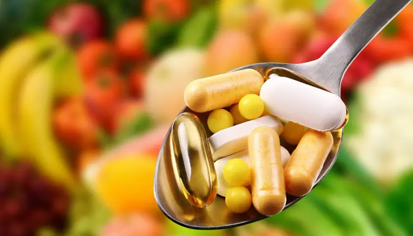 Löffel voller Pillen und Kapseln, die bei Krankheit helfen. Welche Vitamine wenn man krank ist? Bunte Früchte im Hintergrund.