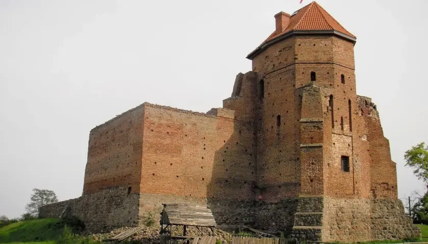Zamek w Liwie, ceglana twierdza z wieżą i dachem, stoi na wzgórzu.