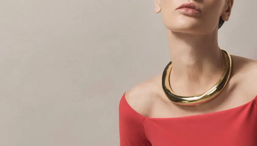A woman wears a bold gold necklace with a curved neckline, complementing her red off-the-shoulder top.