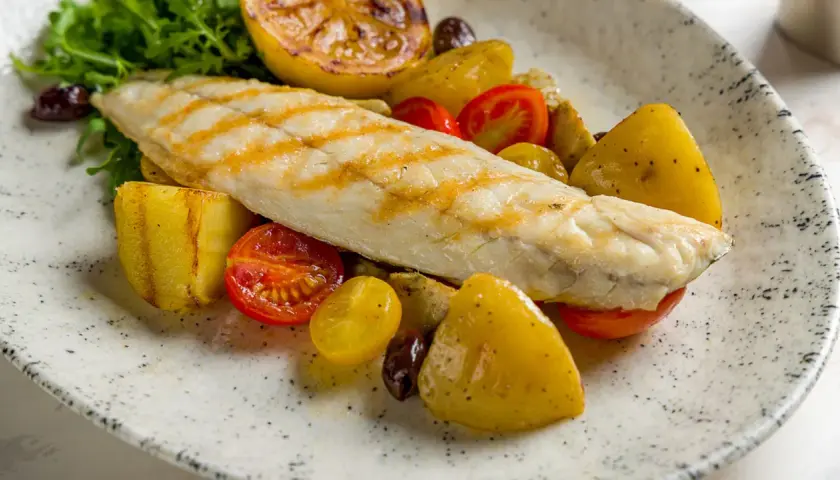 Filetto di persico grigliato su un piatto con patate arrosto, pomodorini, olive e limone caramellato.