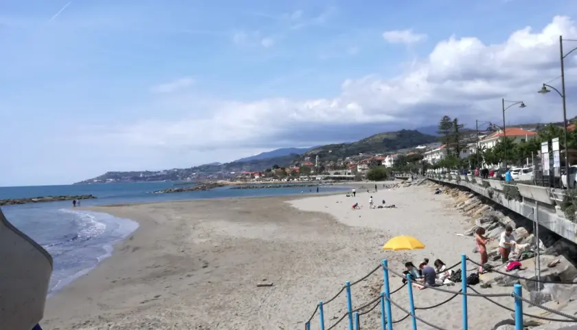 Plage de sable à Santo Stefano al Mare, avec des gens profitant du soleil et de la mer. La ville s'étend le long de la côte, bordée de collines verdoyantes.