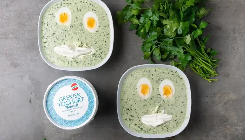 Två skålar med spenatsoppa som ser ut som ansikten, med ägg som ögon och gräddfil som mun. Bredvid står en burk grekisk yoghurt och en grön kvist persilja. Ett roligt spenatsoppa recept!