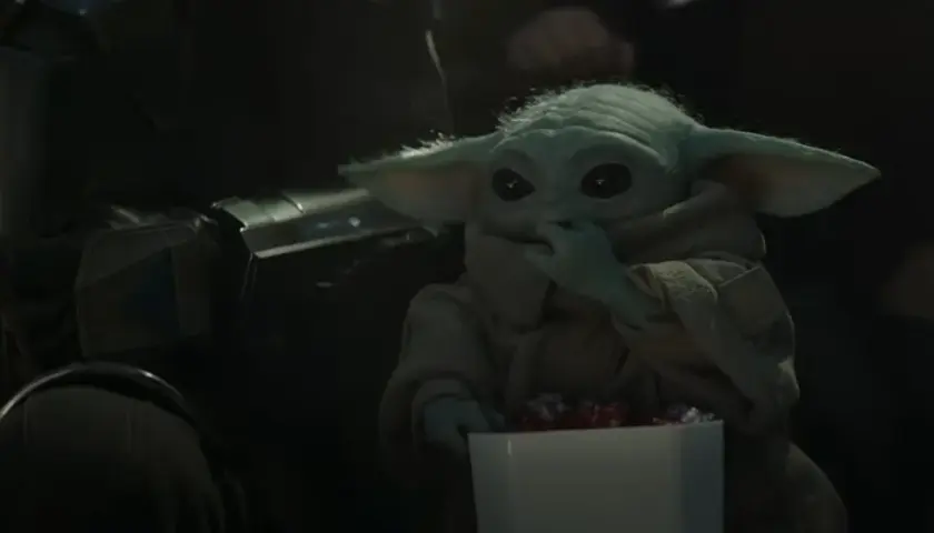 Dziecko z mandalorian film, w brązowym płaszczu, je słodycze z białego pudełka. Obok widać fragment zbroi Mandalorianina.