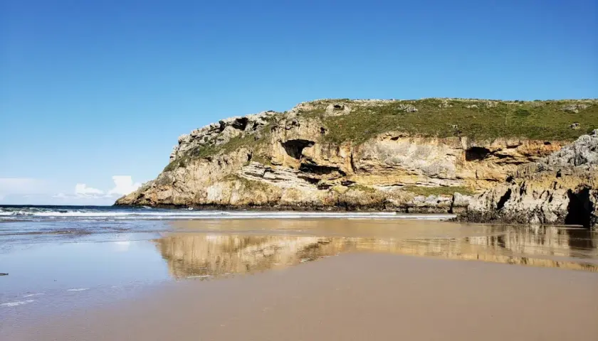 Reflejos dorados en la arena mojada de la playa de Fuentes, con acantilados cubiertos de verde bajo un cielo azul.