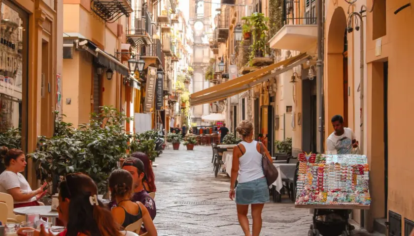 A woman walks down a bustling street in Sicily, past shops and cafes. The scene suggests a vibrant, lively atmosphere, answering the question: is Sicily safe for tourists to enjoy?