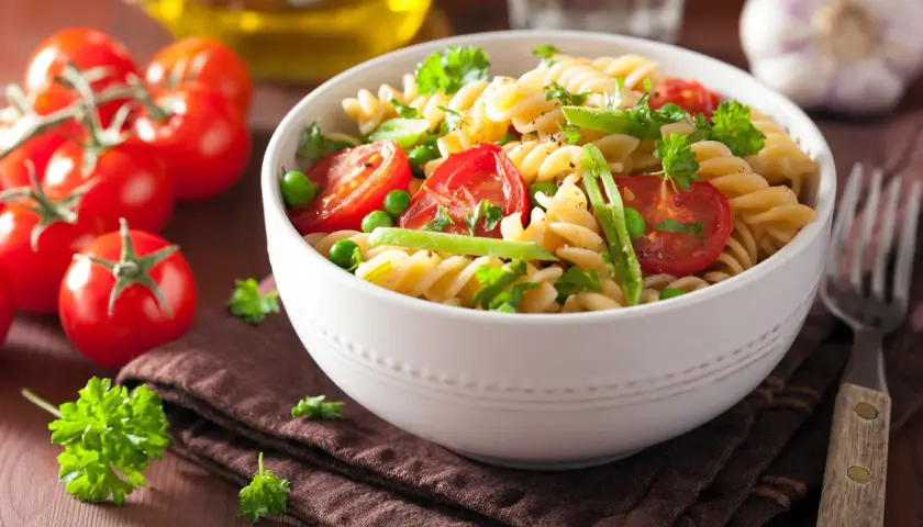 Un delicioso plato de pasta con verduras frescas, como tomates cherry, guisantes y perejil, servido en un cuenco blanco.