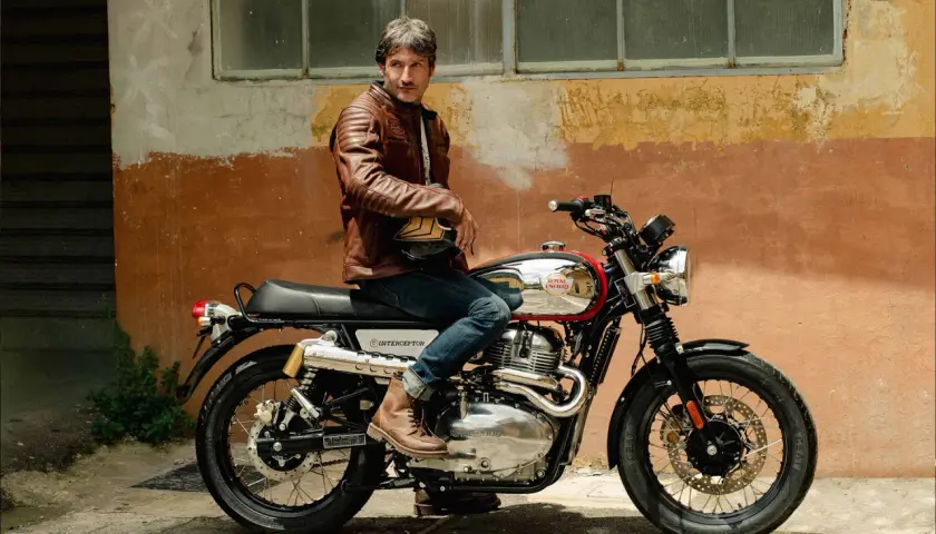 Uomo con giacca in pelle marrone seduto su una moto Royal Enfield Interceptor. I terminali moto cromati brillano sotto il sole.
