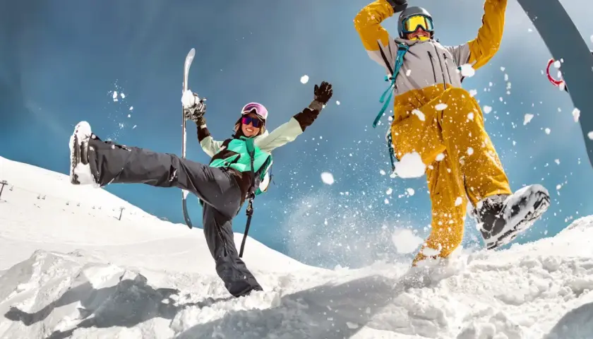 Dwóch narciarzy w akcji na śniegu, jeden z nartami, drugi na snowboardzie, z rozbryzgami śniegu.
