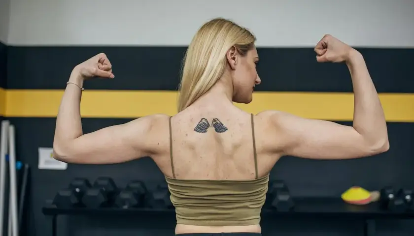 Silna kobieta z tatuażem skrzydeł na plecach napina biceps. Zastanawia się, ile po tatuażu nie można ćwiczyć, by go nie uszkodzić.