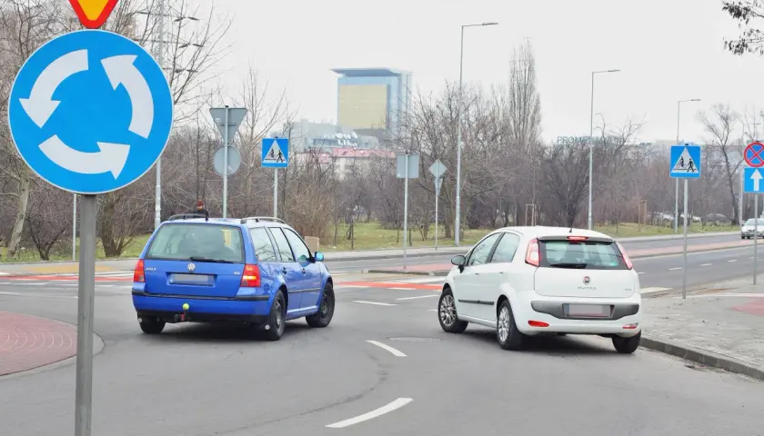 Niebieski Volkswagen i biały Fiat na rondzie. Kierowcy zastanawiają się, który zjazd z ronda na który pas wybrać, by bezpiecznie kontynuować jazdę.