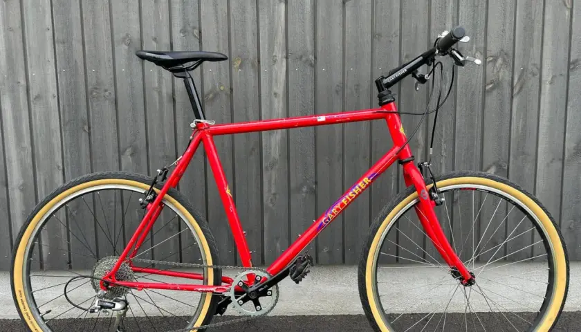 A vintage red Gary Fisher Advance mountain bike with tan tires stands against a wooden fence.