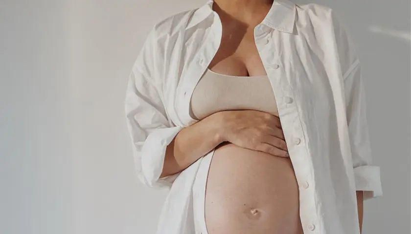 Przyszła mama w białej koszuli czule trzyma brzuch. Zastanawia się nad depilacją laserową w ciąży.