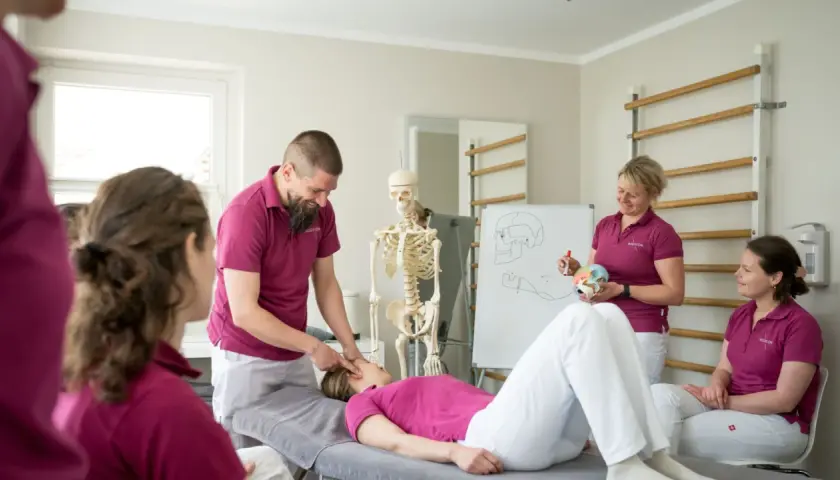 Ein Mann erklärt einer liegenden Frau eine physiotherapeutische Behandlung. Ein Skelett und eine Tafel mit anatomischen Zeichnungen verdeutlichen den Unterschied zwischen Ergo- und Physio-Therapie.