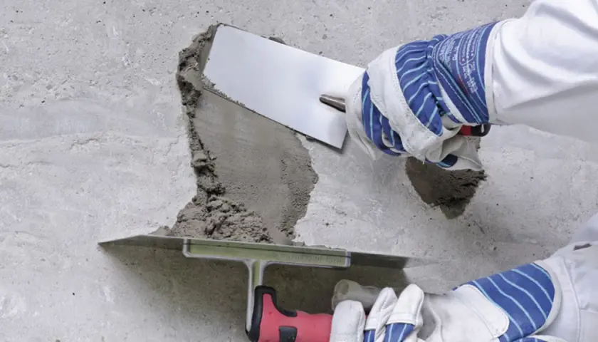 Naprawa betonu na zewnątrz: ręce w rękawicach nakładają zaprawę kielnią na uszkodzoną powierzchnię.