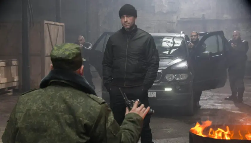 W magazynie, wśród dymu i ognia, mężczyzna w czapce i kurtce bomberce, niczym pitbull, ostatni pies, mierzy z pistoletu do żołnierza.