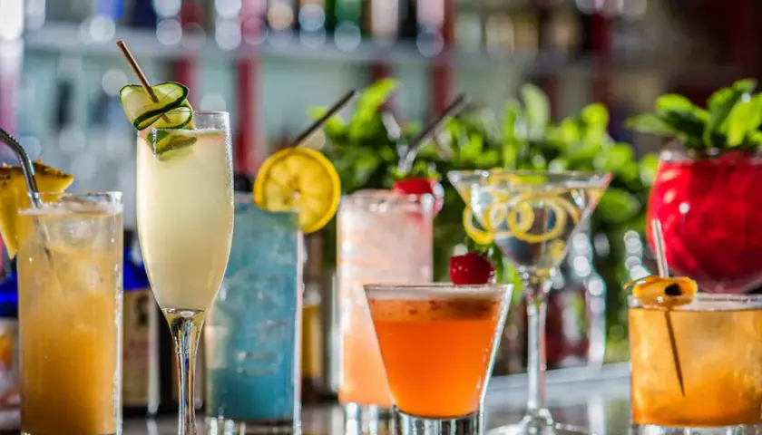 En rad färgglada drinkar, inklusive en sockerlag drink med gurka, står uppradade på en bardisk.