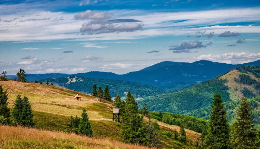 Widok na malowniczą **przełęcz Przegibek** z drewnianą chatką i wieżą widokową na zboczu. W tle gęsto zalesione góry pod błękitnym, chmurnym niebem.