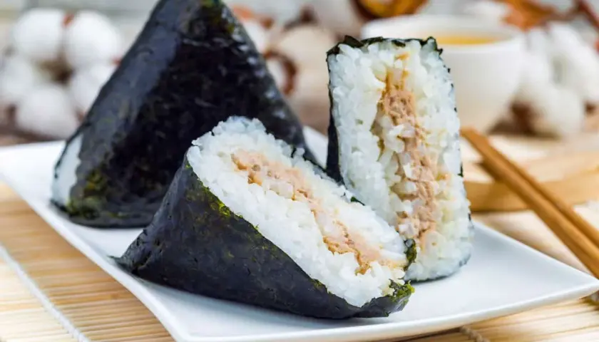 Three triangular onigiri triangle sushi with tuna filling, wrapped in nori seaweed, sit on a white plate with chopsticks and a cup of tea in the background.