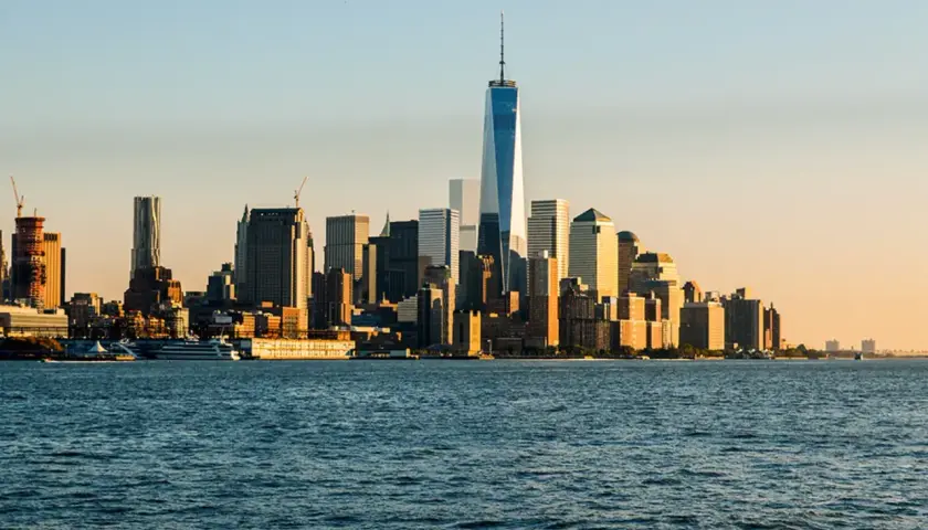 Widok na Manhattan z rzeką w pierwszym planie. W oddali widać drapacze chmur, w tym nowoczesne wieże World Trade Center, oświetlone zachodzącym słońcem.