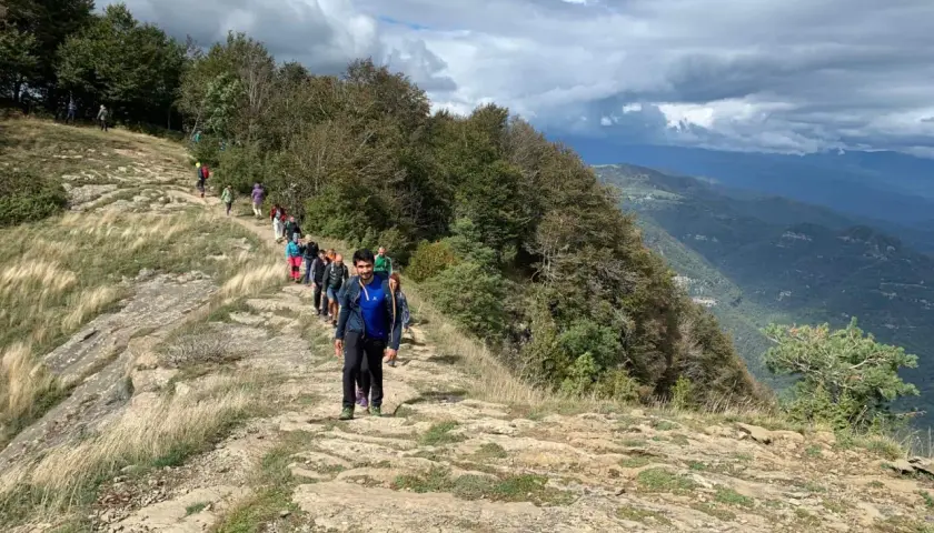Grupo de excursionistas recorre la ruta puigsacalm, un sendero rocoso con vistas a un valle boscoso bajo un cielo nublado.